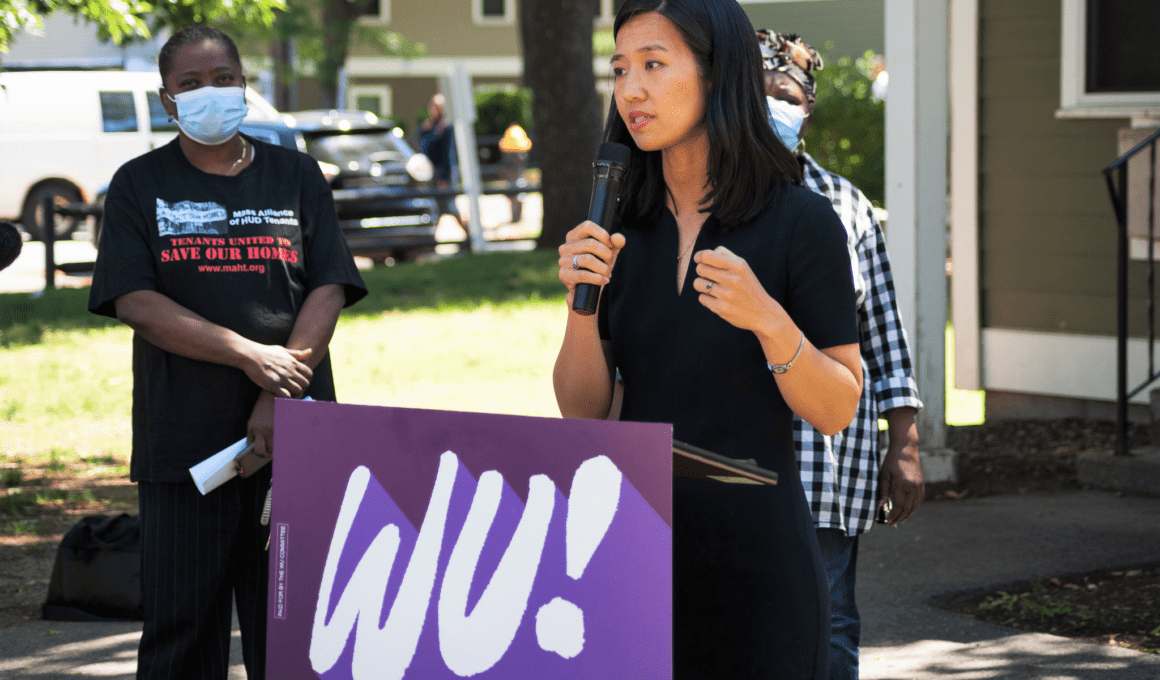 Photo of an Asian woman (Michelle Wu) speaking from a podium in outdoors Boston, with onlookers wearing masks standing behind her
