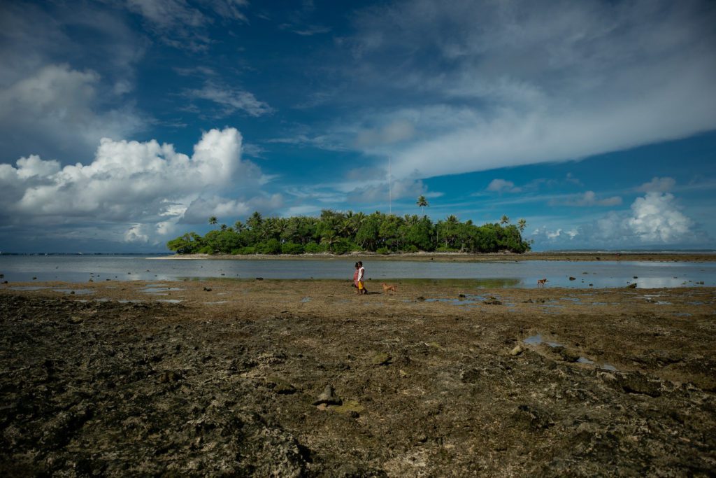 Photo of an island filled with green trees as two Marshallese people walk on a dirt wasteland in the foreground