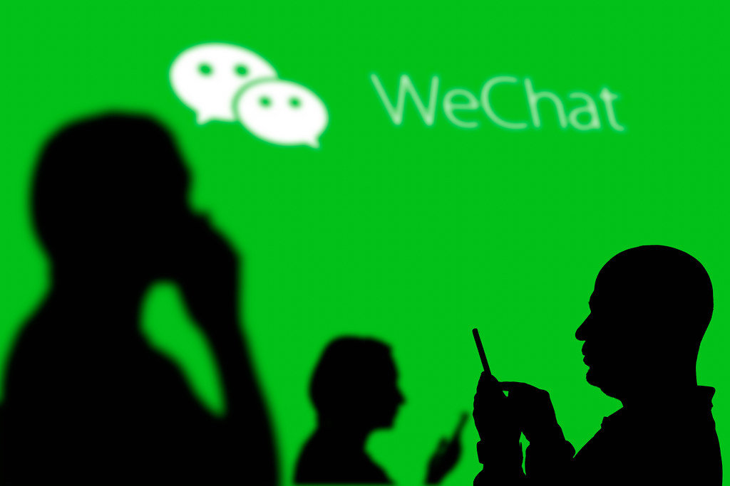 Photo showing silhouettes of people holding phones against a bright green backdrop that displays the logo for WeChat, where misinformation is rampant.