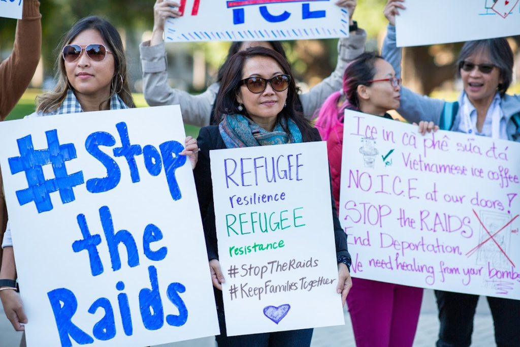 Photo of a group of people holding signs that say "Refugee resilience, refugee resistance," "#StoptheRaidss" and "NO ICE at our doors!" as part of a demonstration against the deportation of Southeast Asian refugees