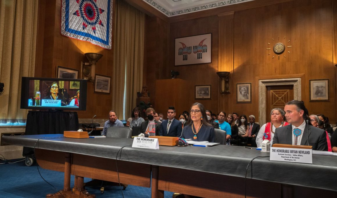 Photo of Deb Haaland sitting at a table in a hearing room as she testifies on the Native American, Alaskan Native, and Native Hawaiian experience in Indian boarding schools.