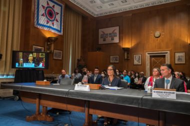 Photo of Deb Haaland sitting at a table in a hearing room as she testifies on the Native American, Alaskan Native, and Native Hawaiian experience in Indian boarding schools.