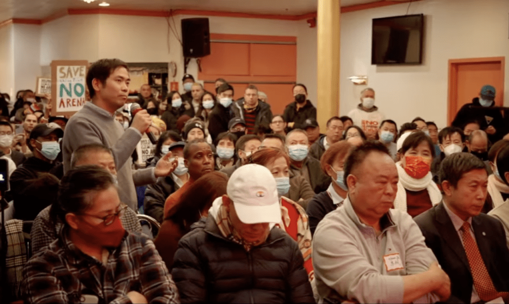 Photo of Philadelphia Chinatown community members at a crowded town hall meeting as a person in the audience speaks into a microphone to ask questions. Some people are holding signs, one of which says "No Arena!"