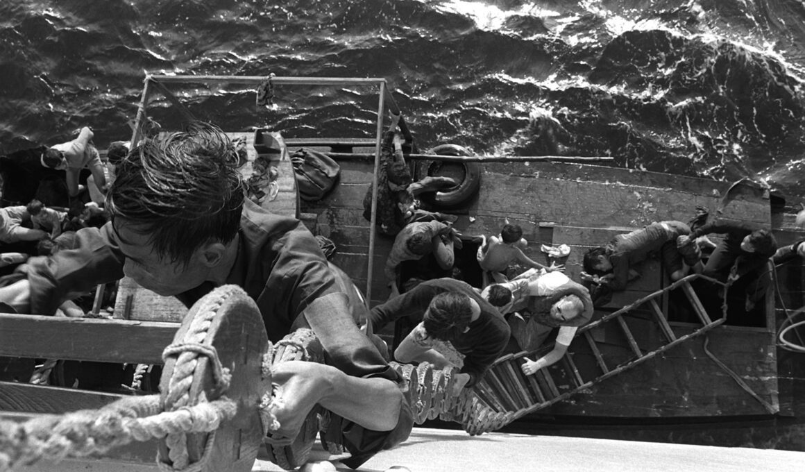 A crewman from the combat stores ship USS SAN JOSE (AFS-7) helps Vietnamese refugees climb from their fishing boat up a Jacobs ladder to the deck of the ship on January 21, 1980. Photo courtesy of The U.S. National Archives and Defense Visual Information Distribution Service.