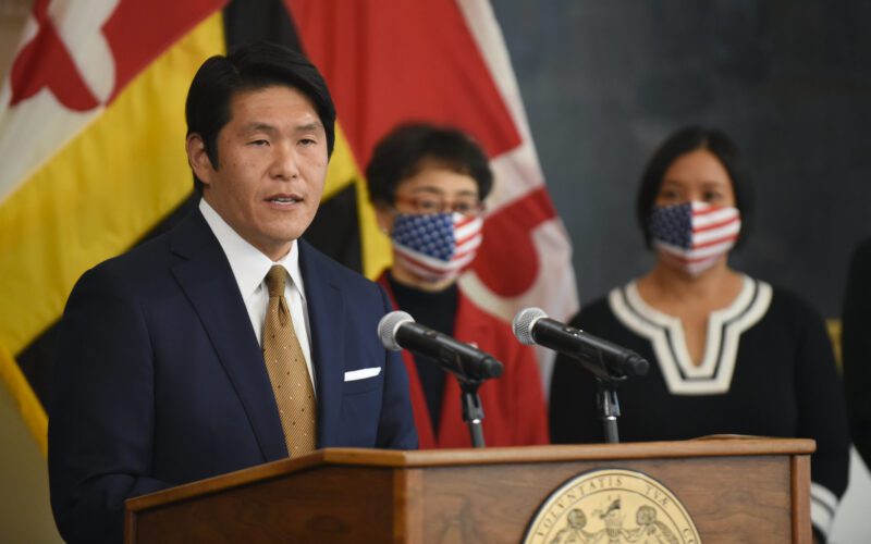 Photo of Robert Hur speaking from a podium as two people, wearing masks, watch from behind.