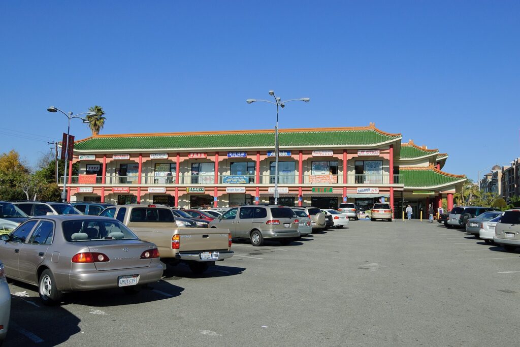 Photo of a shopping plaza with Asian-inspired architecture. Several Asian businesses display signs at the plaza's the front exterior. Cars are parked in the plaza lot.