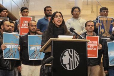 Photo of Kshama Sawant speaking from a podium against caste discrimination as demonstrators stand behind her holding signs.
