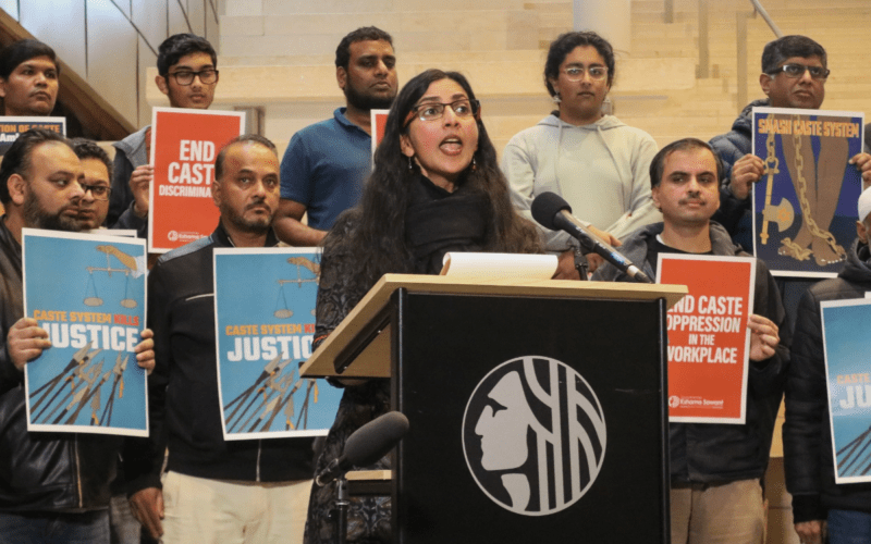 Photo of Kshama Sawant speaking from a podium against caste discrimination as demonstrators stand behind her holding signs.