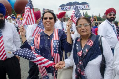 Photo of Sikh Americans waving American flags and signs that say "Sikhs of America" as they march in an Independence Day parade.