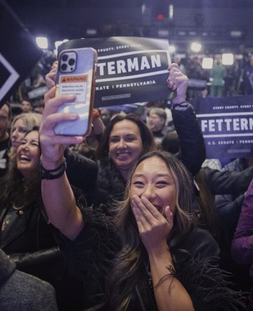 Photo of Annie Wu Henry holding her phone up to the air as she smiles among a crowd of Fetterman supporters