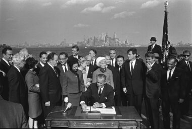 President Lyndon B. Johnson signs the Immigration and Nationality Act of 1965 as people stand around him.