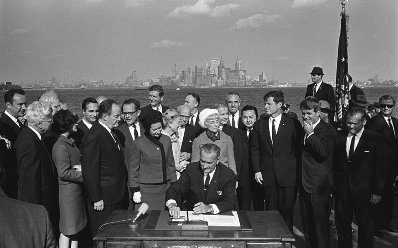 President Lyndon B. Johnson signs the Immigration and Nationality Act of 1965 as people stand around him.