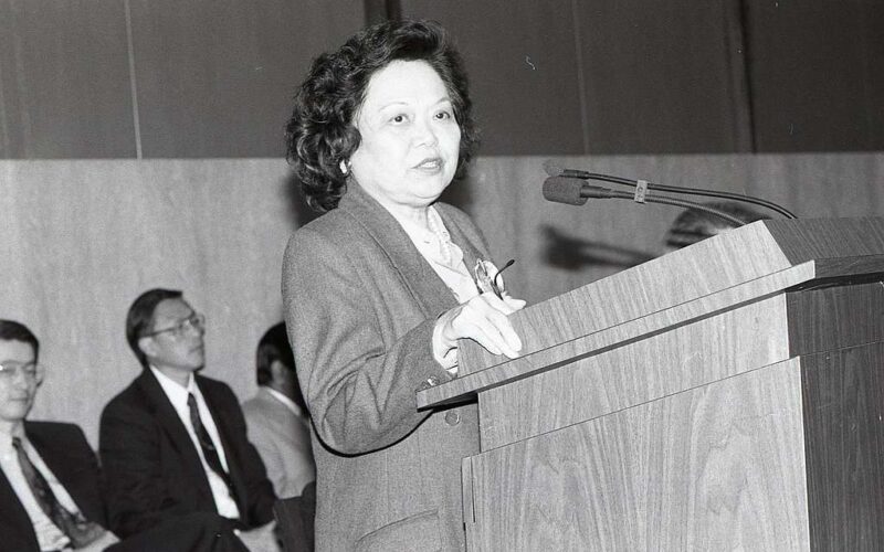Patsy Mink, wearing a suit, standing, speaks at a podium