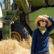 Photo of Mai Nguyen holding on to a hat as they stand in a wheat field in front of a green tractor