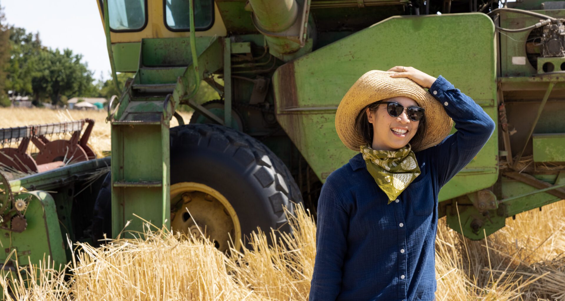 Photo of Mai Nguyen holding on to a hat as they stand in a wheat field in front of a green tractor