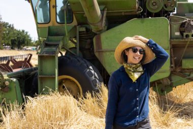 Photo of Mai Nguyen holding on to a hat as they stand in a wheat field in front of a green tractor
